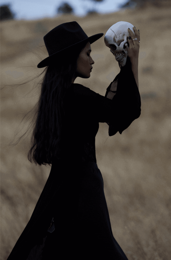 sideview of a women in a black dress. she's holding a skull between her uplifted hands.